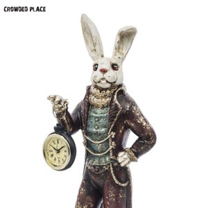 Puede incluir: Una figurita de conejo blanco que lleva un traje marrón y dorado y sostiene un reloj de bolsillo. El conejo tiene ojos rojos y un bigote blanco. El reloj de bolsillo tiene una esfera dorada y agujas negras.