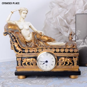 May include: A gold and black clock with a white face and Roman numerals. The clock is on a black base and features a sculpted figure of a woman reclining on a chaise longue.