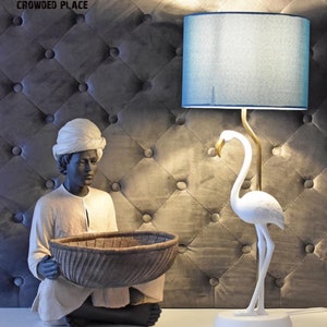 Op de afbeelding: Een decoratieve lamp met een blauwe lampenkap en een goudkleurige, flamingo-vormige voet. Daarnaast een beeld van een persoon met een tulband die een gevlochten mand vasthoudt. De lamp en het beeld staan op een wit oppervlak.