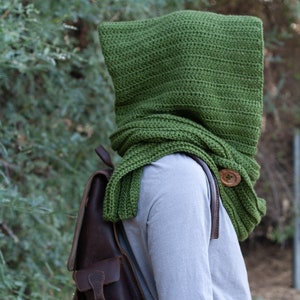 May include: A green crocheted hooded scarf with a wooden button. The scarf is worn over a gray long-sleeved shirt and a brown leather backpack.