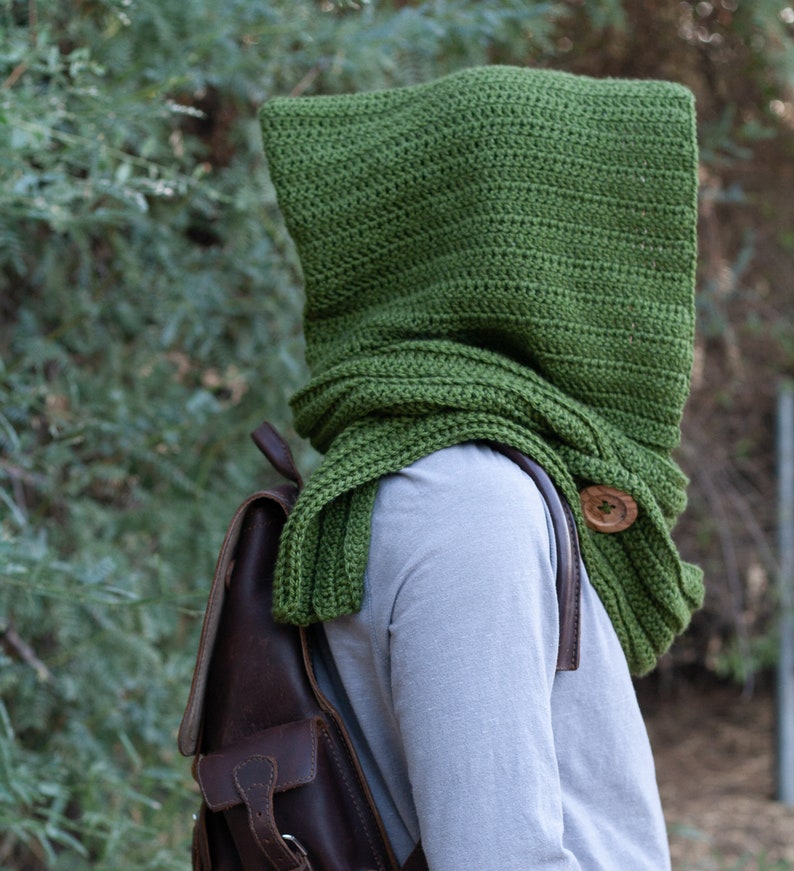May include: A green crocheted hooded scarf with a wooden button. The scarf is worn over a gray long-sleeved shirt and a brown leather backpack.