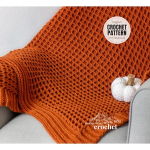 Puede incluir: Una manta de ganchillo de color naranja quemado con un patrón de punto de waffle. La manta está drapeada sobre un sofá gris claro con una calabaza blanca en el sofá. El texto "English Crochet Pattern PDF download" está en un círculo blanco con un borde negro.