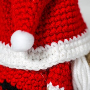 May include: A crocheted Santa Claus ornament with a red hat, white trim, and a white beard. The ornament is made of yarn and has a small white pom-pom on the top of the hat.