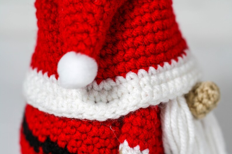 May include: A crocheted Santa Claus ornament with a red hat, white trim, and a white beard. The ornament is made of yarn and has a small white pom-pom on the top of the hat.