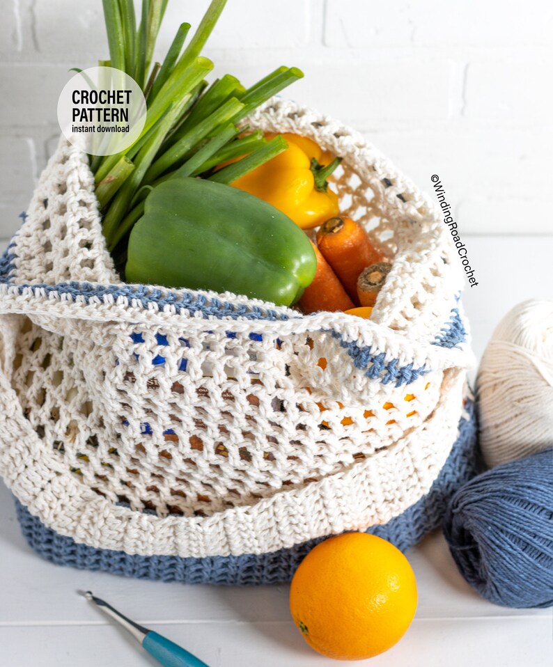 May include: A white crochet mesh bag with blue trim filled with spring onions, yellow and green peppers, carrots, and an orange. The bag is sitting on a white surface with two balls of yarn, one white and one blue.