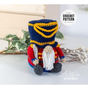 May include: A crocheted gnome doll dressed as a toy soldier. The doll wears a blue hat with yellow trim, a red jacket, and gray pants. The doll has a white beard and holds a gray sword. The text "English CROCHET PATTERN PDF download" is in a circle.