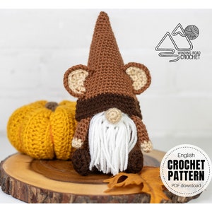 May include: Crochet pattern for a brown and white gnome with a long white beard. The gnome is sitting on a wooden surface with a yellow pumpkin and a brown leaf.