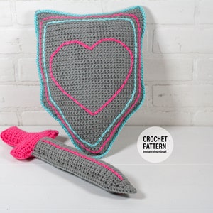 May include: Crochet pattern for a gray heart-shaped shield with blue and pink trim and a gray sword with pink trim. The text "CROCHET PATTERN" and "instant download" is on a white circle.