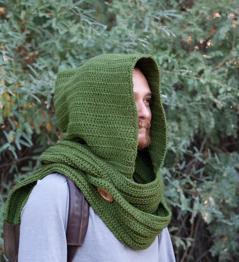 May include: A green crocheted hooded scarf with a wooden button. The scarf is worn by a person with a beard.