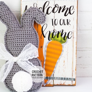 May include: A gray crocheted bunny with a white lace bow and a white pom-pom tail. The bunny is sitting in front of a wooden sign that says "Welcome to our home" with a painted carrot.