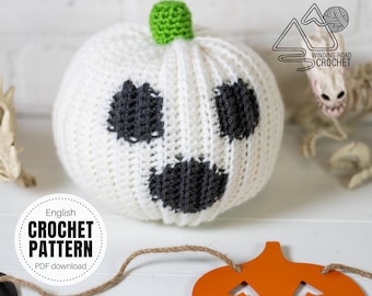 May include: A white crocheted pumpkin with a green stem and black eyes and mouth. The pumpkin is sitting on a white surface with a brown twine and an orange pumpkin cutout.