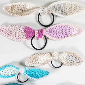 May include: Four crocheted hair scrunchies in different color combinations. The scrunchies are made with a soft, textured yarn and feature a simple bow design. The colors include beige, pink, purple, and blue.