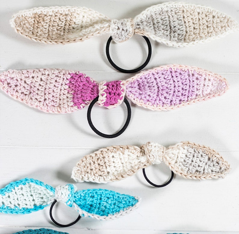 May include: Four crocheted hair scrunchies in different color combinations. The scrunchies are made with a soft, textured yarn and feature a simple bow design. The colors include beige, pink, purple, and blue.