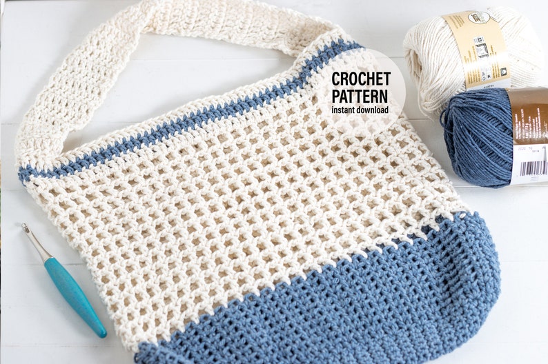 May include: A crocheted tote bag in a white and blue colour scheme. The bag has a wide, open top and a single strap. The bag is made with a mesh stitch and has a solid blue bottom. The bag is lying on a white surface with two balls of yarn in the background.