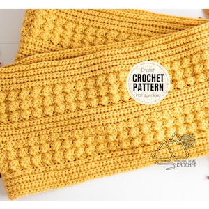 May include: A close-up of a yellow crocheted blanket with a textured stitch pattern. The blanket is folded in half and the edges are visible. The text "English Crochet Pattern PDF download" is visible in a circle on the blanket.