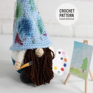 May include: Crochet pattern for a gnome artist holding a paintbrush and a palette. The gnome is wearing a blue hat with a white base and a brown beard. The gnome is standing in front of a small easel with a painting of a tree.
