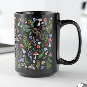 May include: A black ceramic mug with a white and green floral pattern featuring mushrooms, ferns, and a snake. The mug has a white interior and a rounded handle.