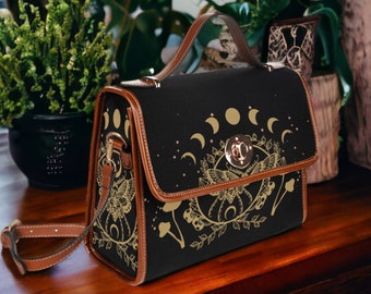 May include: A black canvas shoulder bag with a gold and white moon and butterfly design. The bag has a brown leather strap and a top handle.