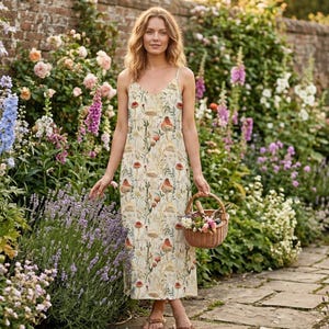 Può includere: Una donna indossa un abito midi color crema con stampa a funghi, spalline sottili e scollo a V. Tiene in mano un piccolo cestino di vimini pieno di fiori in un giardino fiorito, con un sentiero di pietra.