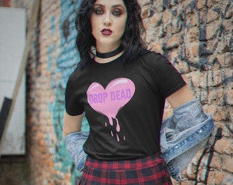 May include: A black t-shirt featuring a pink dripping heart graphic with the words "DROP DEAD" in purple text. The model is wearing a black and red plaid skirt and a denim jacket.