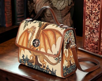 May include: A beige handbag with a dragon illustration. The bag features a brown leather handle, trim, and shoulder strap. The dragon is depicted with orange wings and a long tail. The bag has a silver clasp and is sitting on a wooden table.