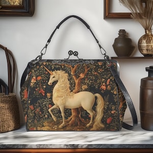 Puede incluir: Un bolso negro con un diseño de unicornio color crema. El bolso presenta un cierre decorativo, un asa superior y una correa para el hombro. El unicornio está sobre un fondo de árbol y flores. El bolso está sobre una superficie de mármol.