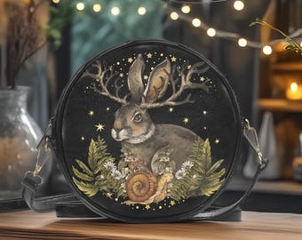 May include: Black round crossbody bag featuring a detailed illustration of a rabbit with antlers, surrounded by stars, ferns, and a snail. The bag has a black strap and gold-colored hardware. The design is set against a dark background.