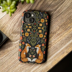 May include: A black phone case featuring a floral design with two foxes facing each other. The case has a glossy finish and is decorated with yellow, orange, and blue flowers. The phone case is on a wooden surface.
