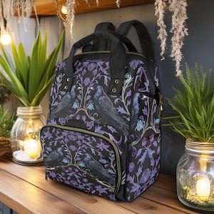 May include: A black backpack with a floral pattern featuring purple and blue flowers and black birds. The backpack has a front pocket with a zipper.