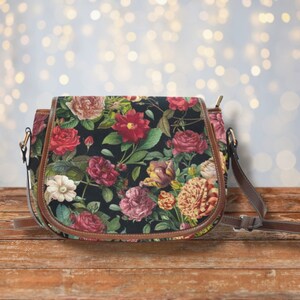 Op de afbeelding: Een bruine leren schoudertas met een bloemenprint met roze, rode en witte bloemen op een zwarte achtergrond. De tas heeft een lange verstelbare schouderband.