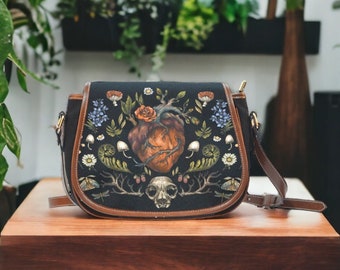 May include: A black canvas satchel bag with a brown leather trim. The bag features a detailed illustration of a heart, flowers, mushrooms, and a skull on a dark background.
