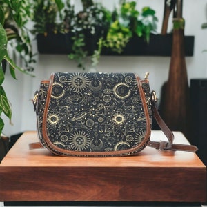 Puede incluir: Un bolso bandolera negro y dorado con un estampado celestial que presenta lunas, estrellas y planetas. El bolso tiene una correa de cuero marrón y un cierre de solapa.