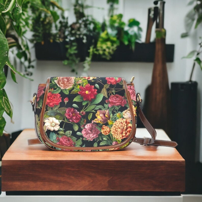 Op de afbeelding: Een zwarte schoudertas met een bloemenprint met rode, roze en witte rozen. De tas heeft een bruine leren afwerking en een lange verstelbare band.