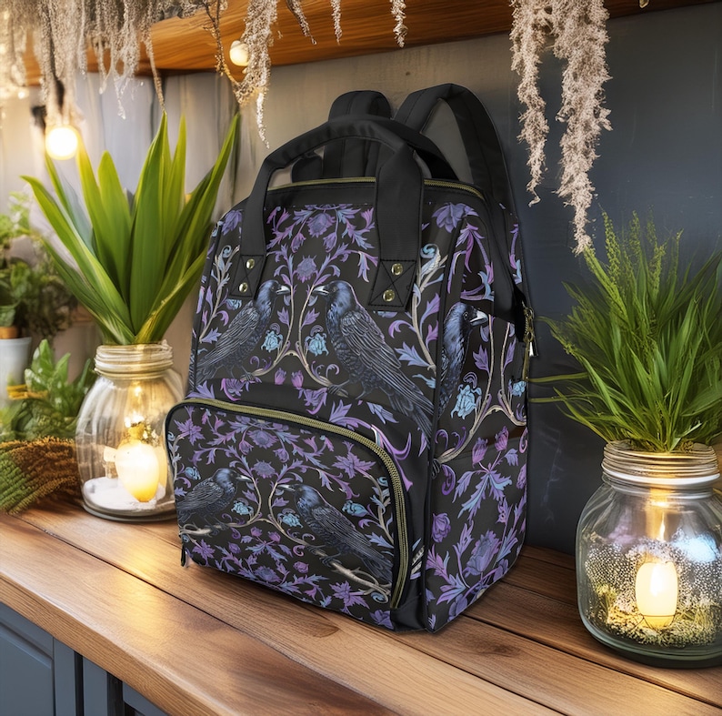 May include: A black backpack with a floral pattern featuring purple and blue flowers and black birds. The backpack has a front pocket with a zipper.