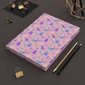 May include: A pink notebook with a repeating pattern of ghosts, bats, skulls, crosses, and other Halloween-themed imagery. The notebook is lying on a gray surface with two pencils and gold paper clips.