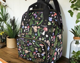 May include: A black backpack with a zippered front pocket. The backpack is covered in a colorful print of mushrooms, ferns, flowers, and other forest elements.