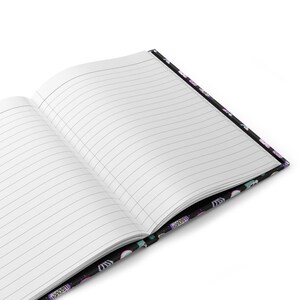 K&ouml;nnte beinhalten: Ein schwarzes Notizbuch mit einem bunten Muster aus Einh&ouml;rnern, Sternen und Regenb&ouml;gen auf dem Einband. Das Notizbuch ist ge&ouml;ffnet und zeigt linierte Seiten.