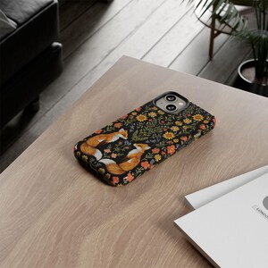 May include: A black phone case featuring a floral pattern with orange, yellow, and red flowers. Two foxes are depicted in the center, facing each other. The case is on a light brown wooden surface.