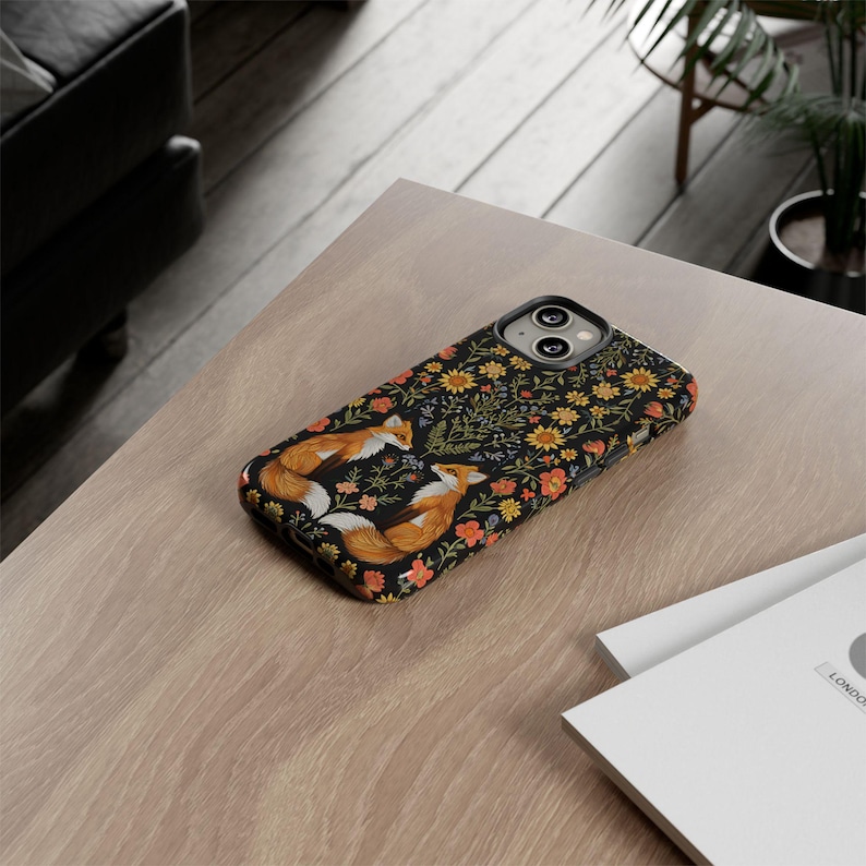 May include: A black phone case featuring a floral pattern with orange, yellow, and red flowers. Two foxes are depicted in the center, facing each other. The case is on a light brown wooden surface.