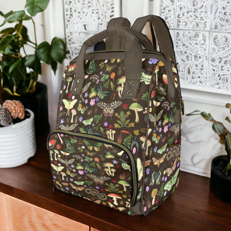 Op de afbeelding: Een bruine rugzak met een kleurrijk patroon van vlinders, paddenstoelen en bloemen. De rugzak heeft een handvat aan de bovenkant, verstelbare bandjes en een voorvak met ritssluiting. De tas is ongeveer 40 cm hoog, 30 cm breed en 15 cm diep.