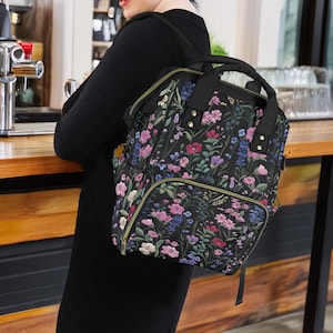 Könnte beinhalten: Ein schwarzer Rucksack mit floralem Muster, goldenen Akzenten und einer Reißverschlusstasche. Der Rucksack hat verstellbare Träger und einen Tragegriff oben.
