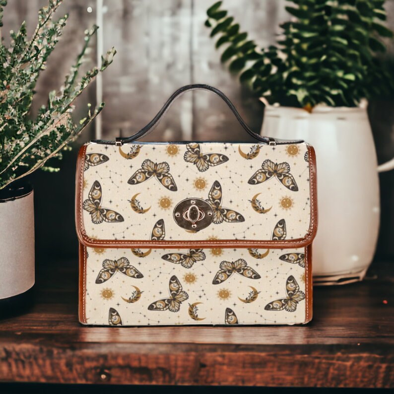 May include: A brown leather satchel with a cream-colored fabric front featuring a repeating pattern of gold butterflies, moons, and stars. The bag has a top handle and a clasp closure.