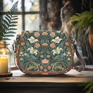Peut inclure: Sac à bandoulière à motif floral avec un fond vert foncé, orné de lys blancs, de fleurs couleur pêche et de feuilles vertes. Le sac a une bordure et une sangle en cuir marron, posé sur une surface en bois.
