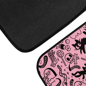 May include: Pink and black patterned doormat with a skull, cat, and other spooky designs. The text "GRRRRR", "RIP", "BOO000", and "ZZZ" are included in the design.