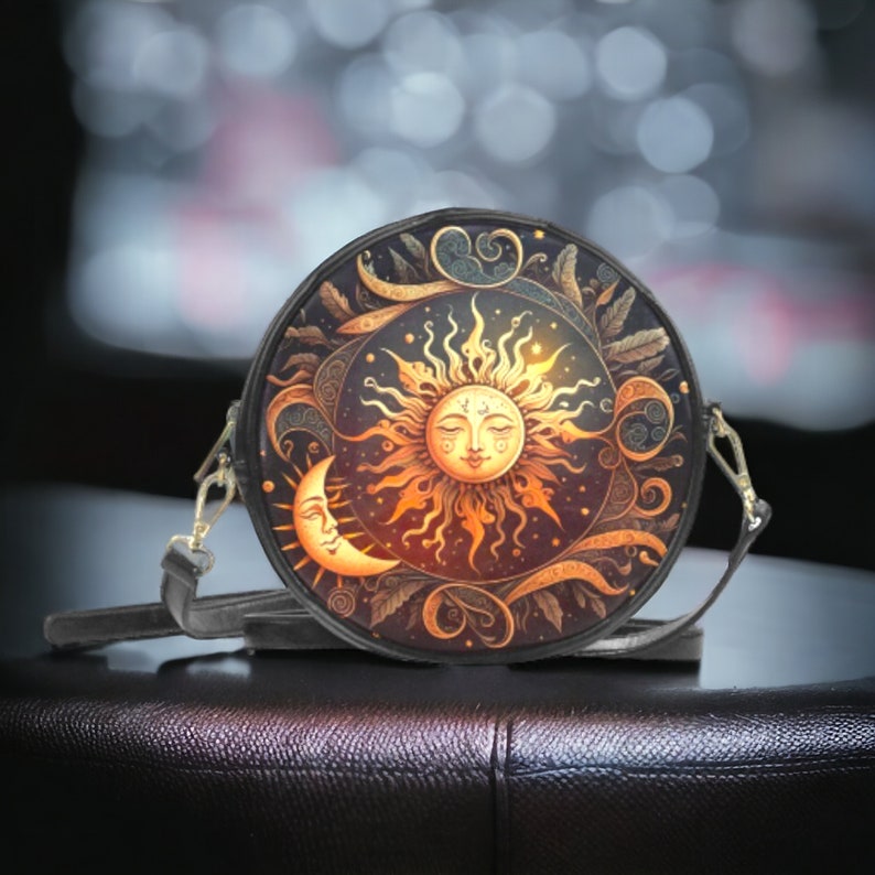 Puede incluir: Bolso bandolera redondo negro con un dise&ntilde;o de sol y luna. El sol es dorado con una cara sonriente, y la luna tiene forma de media luna. El bolso tiene una correa larga y ajustable y herrajes dorados.
