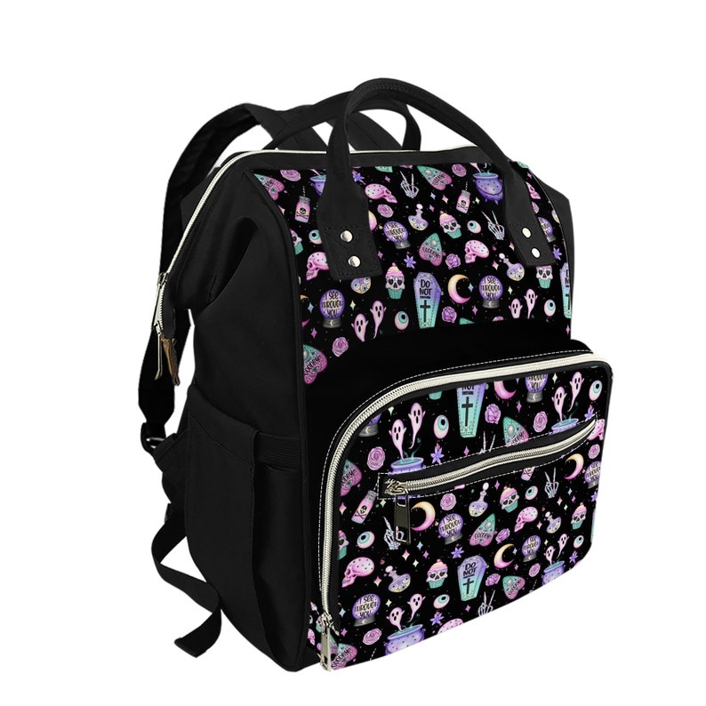 May include: A black diaper bag with a whimsical, gothic-inspired print featuring ghosts, eyeballs, moons, and other spooky elements in pastel colors. The bag has multiple pockets and a zippered closure.