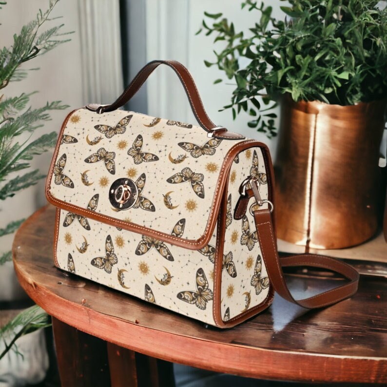 May include: A beige satchel bag with a brown leather strap and handle. The bag features a repeating pattern of butterflies, moons, and suns on a white background.