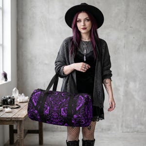 May include: A black duffel bag with a purple gothic pattern featuring moths, moons, and floral designs. The bag has black handles and a zipper closure. The bag is held by a person wearing a black hat and a gray cardigan.