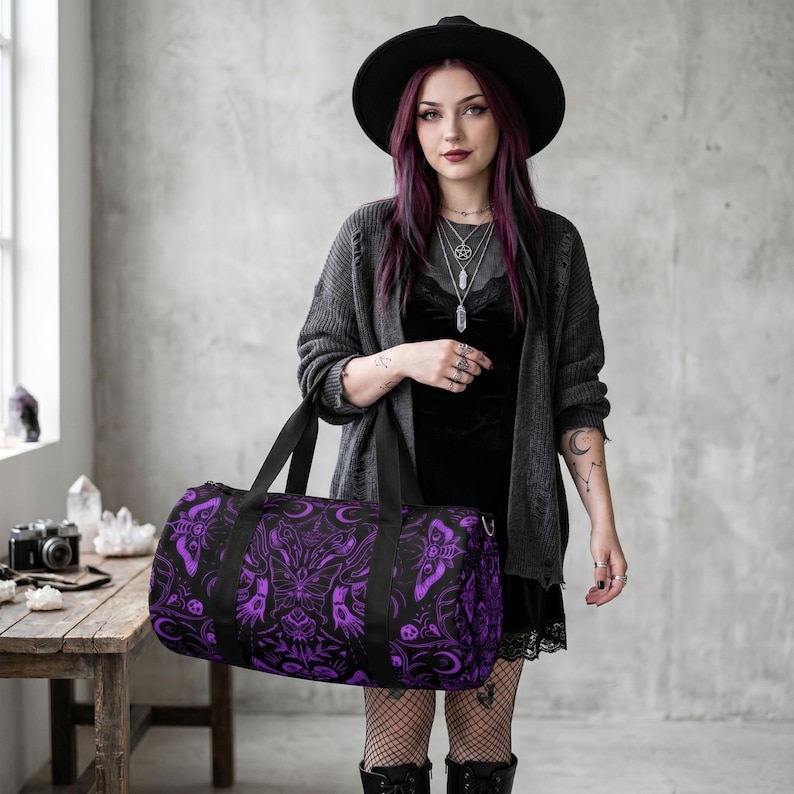 May include: A black duffel bag with a purple gothic pattern featuring moths, moons, and floral designs. The bag has black handles and a zipper closure. The bag is held by a person wearing a black hat and a gray cardigan.