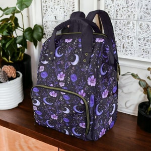 Può includere: Uno zaino nero con un motivo floreale con fiori viola, lune crescenti e stelle. Lo zaino ha una tasca frontale con cerniera e spallacci regolabili.
